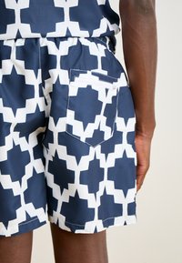 Marineblaue Schwimmshorts mit einem weißen geometrischen Muster. Verfügt über einen elastischen Bund und eine Gesäßtasche mit Reißverschluss. Glatte Stofftextur.