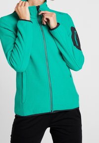 Fleecejack met een heldere teal kleur, een rits aan de voorkant, hoge kraag en zwarte accenten op de schouders. Getextureerde stof met zijzakken.