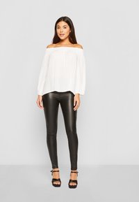 Witte off-the-shoulder blouse met elastische halslijn, gecombineerd met zwarte glanzende leren legging en zwarte strappy hakken, staande tegen een grijze achtergrond.