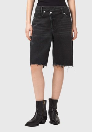 Nő fekete, magas derekú, nyers szegélyű farmer bermuda shortot és fekete bokacsizmát visel oldalsó cipzárral, szürke háttér előtt áll.