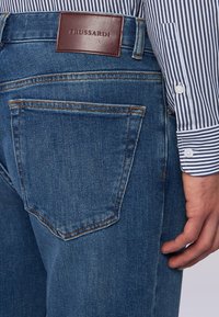 Jeans in denim blu con una finitura testurizzata, caratterizzati da un'etichetta in pelle marrone con il logo TRUSSARDI e tasche posteriori tradizionali. Camicia a righe visibile.