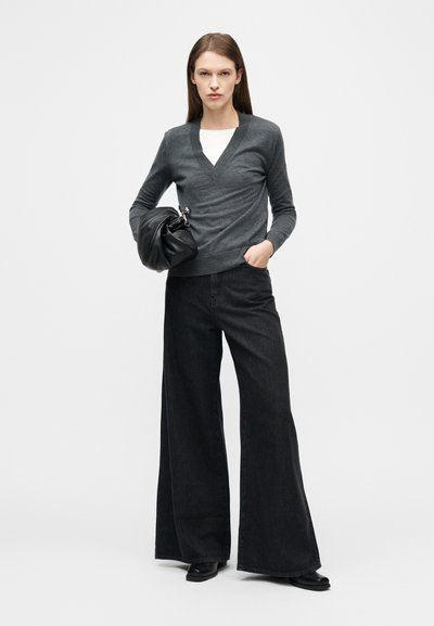 Γυναίκα που στέκεται μπροστά σε λευκό φόντο, φοράει σκούρο γκρι πουλόβερ V-neck, μαύρο παντελόνι με φαρδιά μπατζάκια, μαύρα μποτάκια και κρατά μαύρο τσαντάκι χειρός.