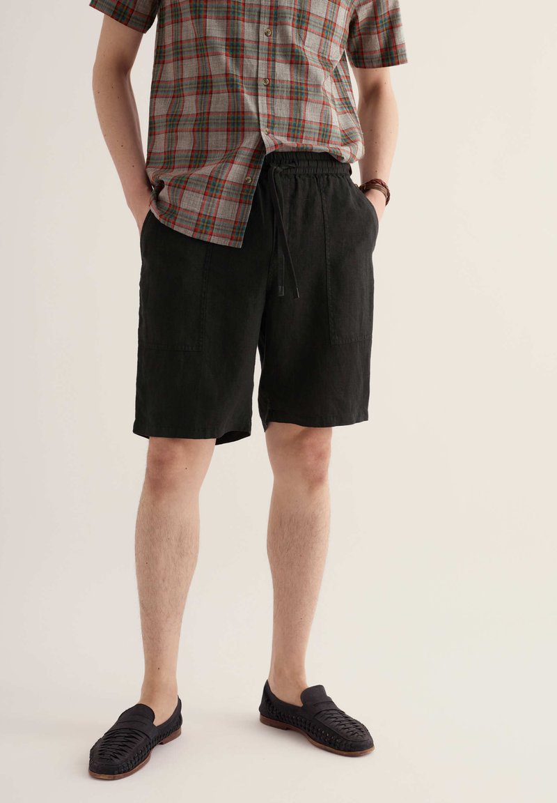 Personne portant un short noir à cordon, une chemise à manches courtes à carreaux et des chaussures noires tissées sans lacets, debout les mains dans les poches.