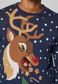 Gestrickter Pullover mit braunem Rentier-Design, Geweihen, roter Nase und festlichen Elementen wie Zuckerstangen und Sternen vor einem marineblauen Hintergrund.