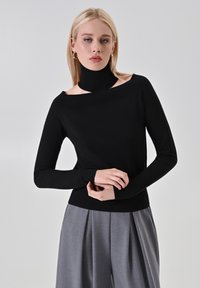 Maglione nero a maglia con colletto alto e design scollato. Maniche lunghe e corpo aderente, abbinato a pantaloni grigi a pieghe.