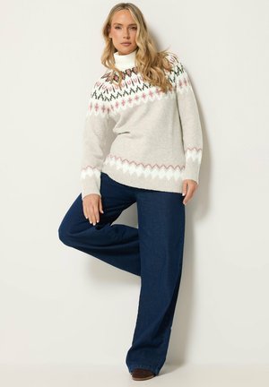 Femme aux longs cheveux blonds portant un pull blanc à motifs, un col roulé blanc, un pantalon large bleu foncé et des chaussures marron, appuyée contre un mur.