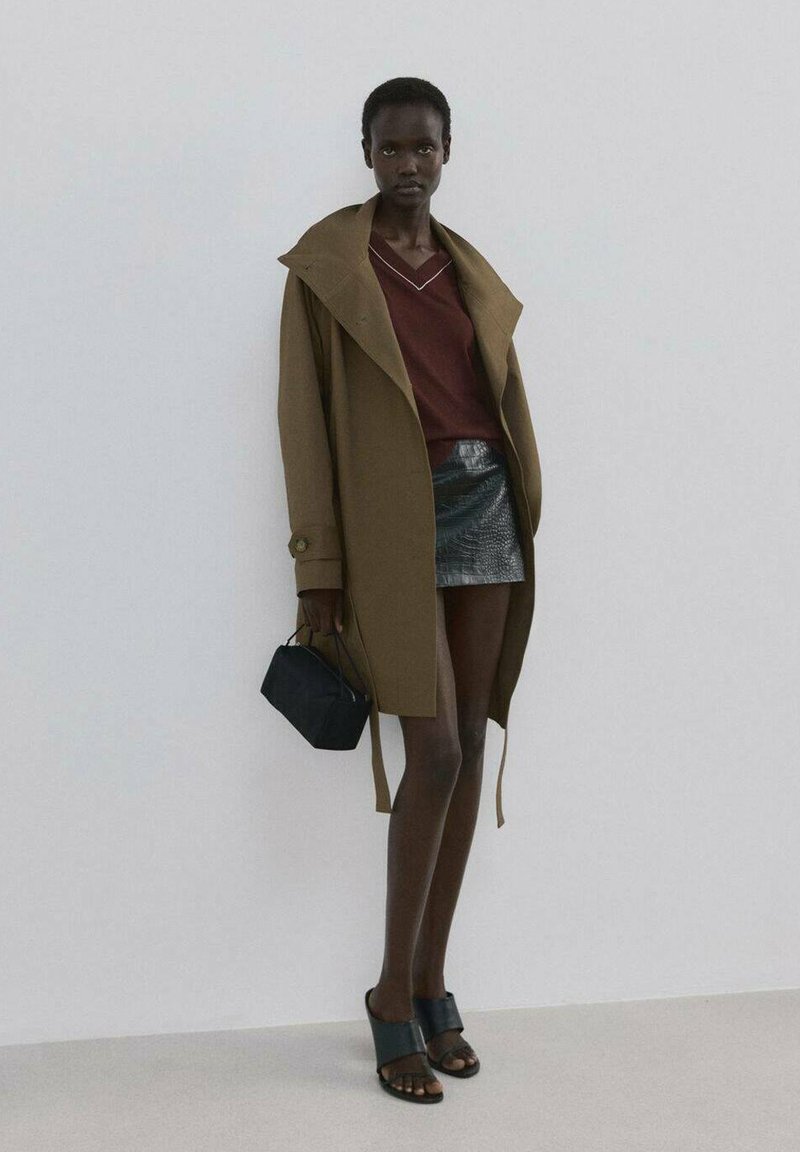 Manteau oversize vert olive ; haut bordeaux avec couture blanche ; mini-jupe noire brillante ; sac à main noir ; sandales noires à larges brides.