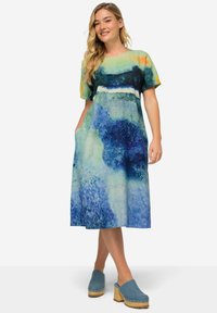 Kurzärmliges, knielanges Kleid mit einem blau-grünen abstrakten Muster, aus leichtem Stoff mit Taschen. Kombiniert mit hellblauen Clogs.