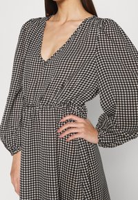 Svartvitt houndstooth-mönstrat klänning med djup V-ringning, puffärmar och åtstramad midja, gjord av lättviktsmaterial.
