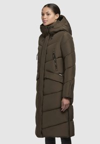 Giacca a vento lunga, di colore verde oliva, imbottita e con cappuccio, caratterizzata da un design trapuntato diagonale, tasche con zip e cordini regolabili.