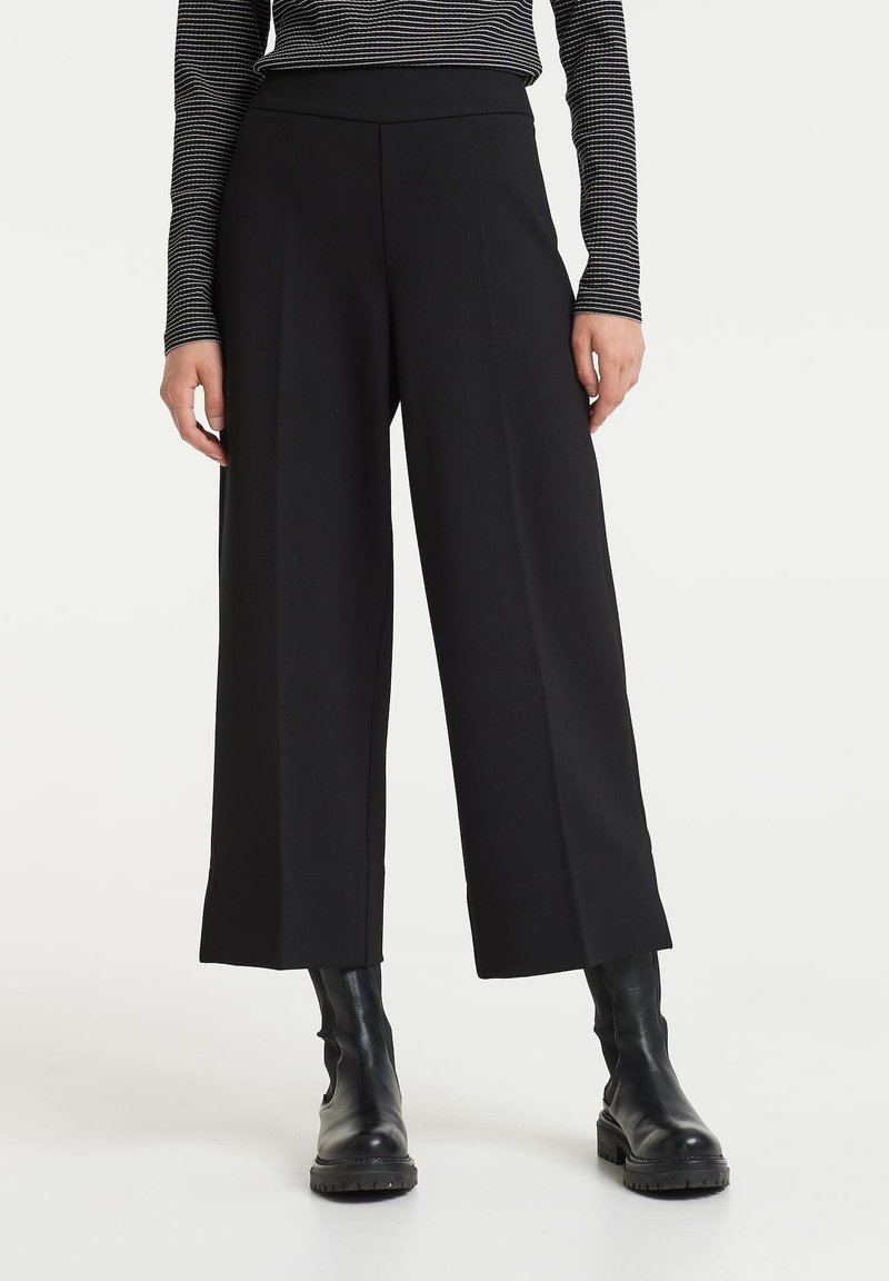 Pantalons larges noirs en tissu lisse ; taille haute, plis nets et coupe droite. Associés à des bottines noires.