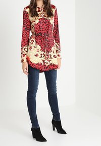Bluse med rød og sort leopardmønster samt gyldent blomsterdesign, lange ærmer, knapnedlukning og justerbar snor i taljen, parret med mørke jeans og sorte ankelstøvler.