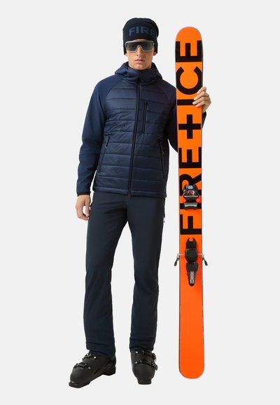 Marineblaue isolierte Jacke, schwarze Hose, schwarze Skischuhe und ein orangefarbener Ski mit "FIRE+ICE" Branding, der aufrecht gehalten wird. Inklusive schwarzer Mütze und Sonnenbrille.