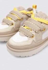 Zapatillas de ante beige con acentos blancos, que presentan tejido texturizado, cordones trenzados beige y correas amarillas brillantes para un detalle adicional.
