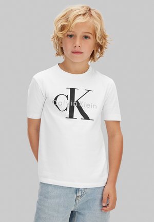 Ξανθό παιδί που φοράει λευκό t-shirt Calvin Klein και ανοιχτόχρωμα μπλε τζιν, όρθιο μπροστά σε απλό γκρι φόντο.
