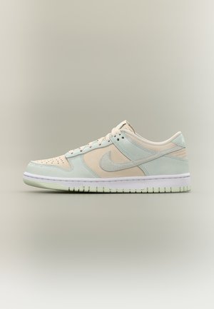 Baskets basses avec panneaux beige et bleu clair, logo Nike swoosh bleu clair, semelle blanche et lacets beige sur un fond neutre uni.