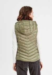 Gilet matelassé vert olive avec capuche, présentant un motif en chevrons, en matériau léger et avec un design à col montant.