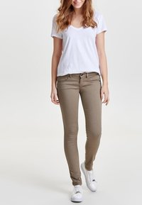 Jeans ajustados beige con bolsillos frontales, passadores para cinturón y cremallera, combinados con una camiseta blanca de cuello en V y zapatillas blancas.