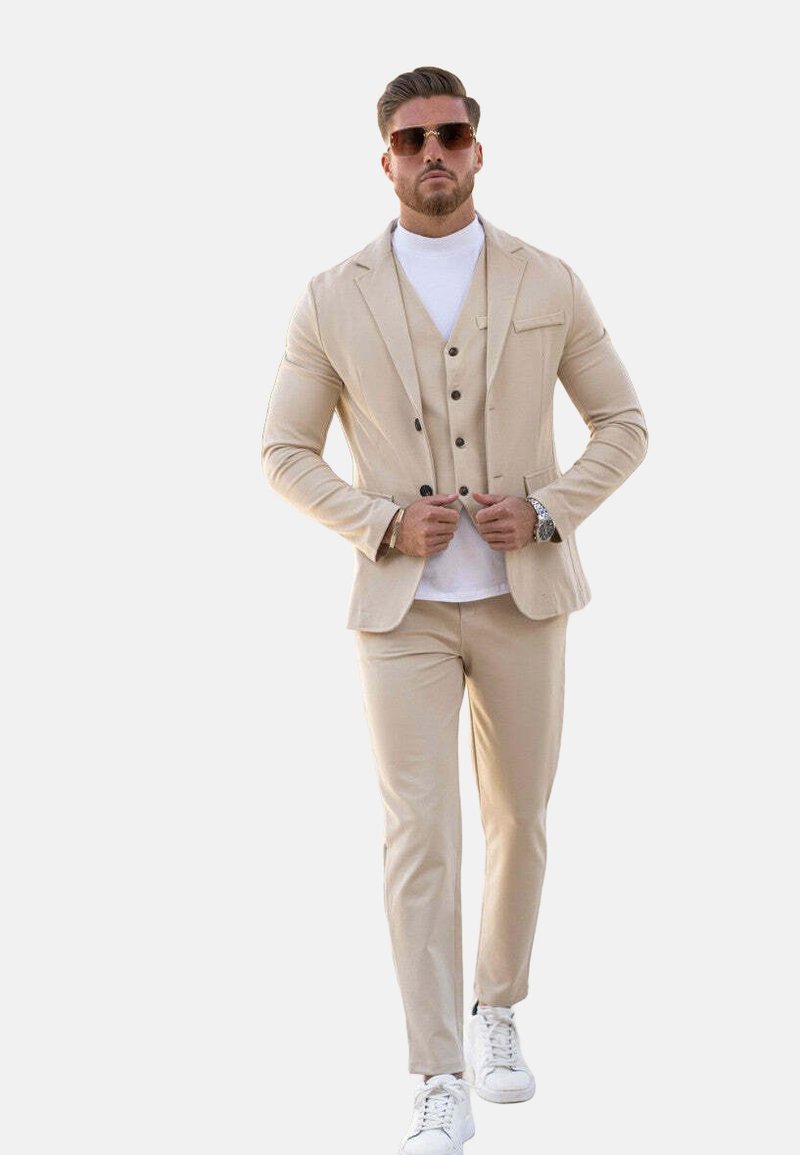 Costume beige avec une veste ajustée et un pantalon assorti, superposé sur un col roulé blanc, associé à des baskets blanches et des lunettes de soleil.