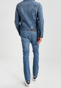 Modrá džínová bunda s lehkým vyblednutím, oprýskanými detaily a kontrastním šitím. Kombinováno s odpovídajícími modrými džínami a bílými teniskami.