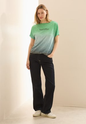 Camiseta verde degradada con el texto "il paradiso", combinada con unos jeans oscuros. La camiseta tiene un corte amplio y una textura suave. Zapatillas blancas casuales.