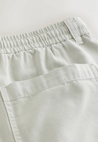 Pantalon gris clair avec ceinture élastique et passants pour ceinture au-dessus d'une grande poche arrière, montrant la texture du tissu et les détails de la couture.