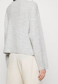 Persona che indossa un maglione corto grigio chiaro e pantaloni color crema, mostrata di spalle su uno sfondo leggero e uniforme.