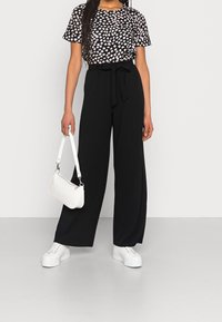 Mujer vistiendo pantalones de pierna ancha negros con un cinturón atado, una blusa con estampado en blanco y negro, zapatillas blancas y sosteniendo un pequeño bolso blanco.