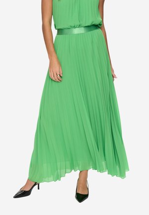 Kvinde iført en grøn plisseret kjole hele vejen ned med matchende bælte og sorte spidse høje hæle, stående mod en hvid baggrund.