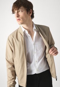 Giubbotto bomber beige con finitura testurizzata e zip frontale, indossato sopra una camicia bianca con bottoni, su uno sfondo neutro.