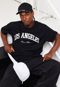 Sort t-shirt med "LOS ANGELES" i hvid, parret med løse mørke jeans. En hvid talje taske sidder mod hoften, med en lynlåsdetalje.