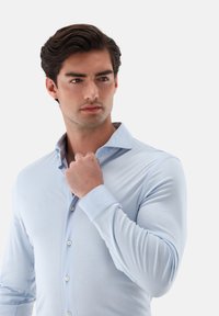 Hellblaues Hemd aus glattem Stoff mit Knopfleiste. Verfügt über einen Stehkragen und taillierte lange Ärmel mit Knöpfen entlang der Vorderseite.