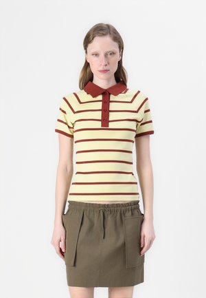 Kvinne iført en kortermet polo skjorte med gule og ruststriper, rustfarget krage og knapper, sammen med et olivengrønt skjørt med store lommer.