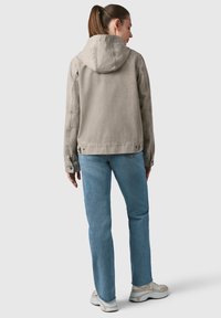 Giacca a cappuccio grigia chiara con una texture liscia, polsini con bottoni e un fit casual, abbinata a jeans azzurri e sneakers beige.