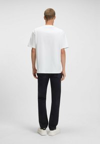 T-shirt bianca in cotone con maniche corte e scollo rotondo, abbinata a jeans neri e sneakers bianche, vista da dietro. Nessun motivo visibile.