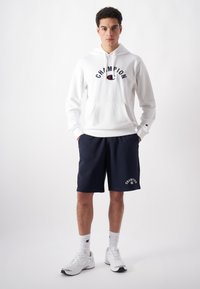 White hoodie with "CHAMPION" logo, navy shorts with logo accent, and white athletic shoes. Casual sport outfit. Smooth cotton fabric.