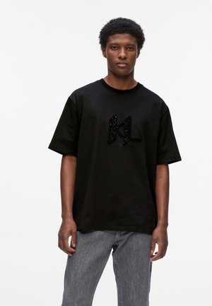 Schwarzes Baumwoll-T-Shirt mit weiten Ärmeln. Hat ein strukturiertes schwarzes "KL"-Logo auf der Vorderseite. Kombiniert mit grauen Jeans.