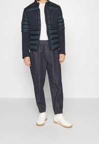 Marinblå vadderad jacka med platta sömmar och fickor med dragkedja, kombinerad med löst sittande mörk denimjoggingbyxor och vita sneakers med beige sula.