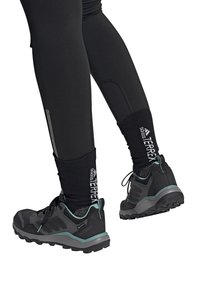 Czarne buty do wędrówek z syntetycznej cholewki i gumowej podeszwy. Wyraźne akcenty w kolorze teal i logo; zestawione z czarnymi legginsami i wełnianymi skarpetami.