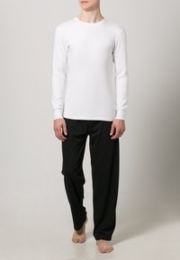 Top branco de mangas longas, com gola redonda, e calças pretas folgadas. Tecido leve com uma textura suave. Pés descalços.