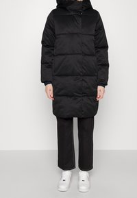 Person med en svart vadderad knälång jacka, svarta byxor med vit sömnad och vita sneakers, stående mot en enkel bakgrund.