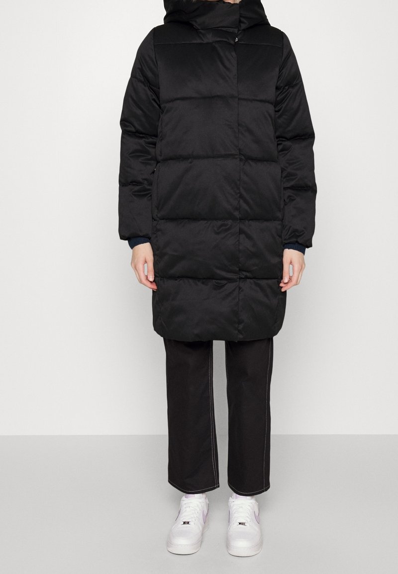 Person med en svart vadderad knälång jacka, svarta byxor med vit sömnad och vita sneakers, stående mot en enkel bakgrund.