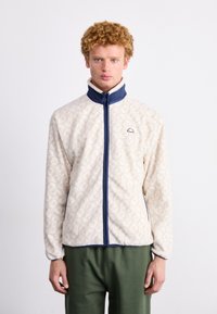 Chaqueta de forro polar en color crema con un ligero patrón, cuello azul marino y detalles en cremallera, con bolsillos y un corte relajado.