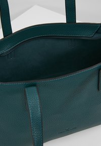 Vihreä tekstuuri nahkainen olkalaukku, jossa on kaksi hihnaa, avoimen ylimmän osan ja sisäisen vetoketjullisen taskun esittely yksinkertaista taustaa vasten.