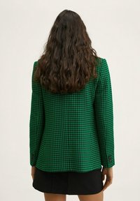 Veste blazer vert à motif pied-de-poule, coupe ajustée, épaules structurées et poignets à boutons, associée à une mini-jupe noire.