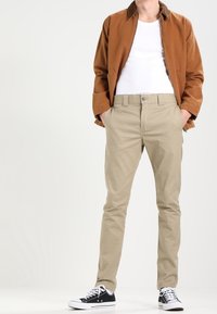 Béžové chinos s úzkým střihem, v kombinaci s bílým tričkem a hnědou bombardérkou, a čern é tenisky s bílou podrážkou a tkaničkami.