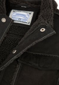 Schwarze Jacke mit weichem, flauschigem Innenfutter und Shearling-Kragen. Zwei Druckknöpfe und ein innen sichtbares Etikett. Strukturierter Stoffdesign.