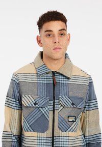 Veste en flanelle à carreaux beige et bleu avec une fermeture éclair intégrale, deux poches avant et une étiquette. Présente une texture douce et un design de col.