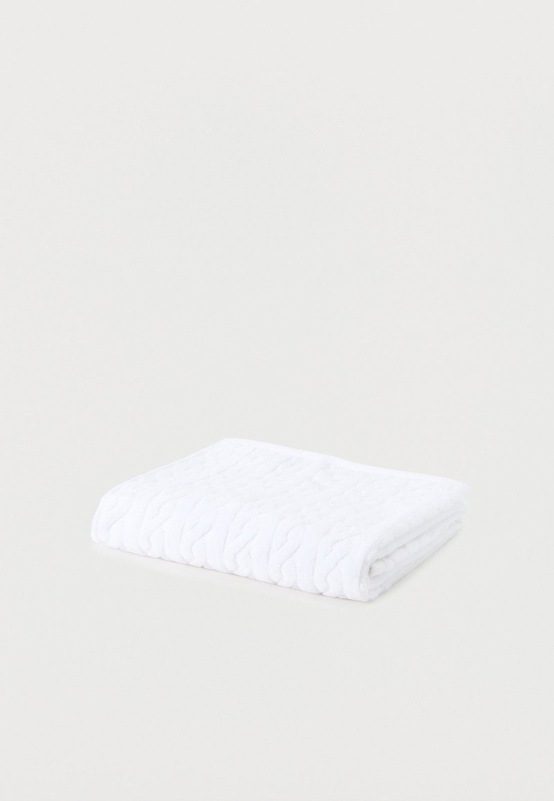 Διπλωμένη λευκή πλεκτή κουβέρτα με πλεγμένο σχέδιο σε απλό ανοιχτό γκρι φόντο.