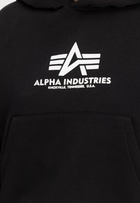 Čierna mikina s kapucňou vyrobená z mäkkej tkaniny s prednou vreckom. Na hrudníku je biele grafické logo s štýlovým textom a dizajnovými detailmi.